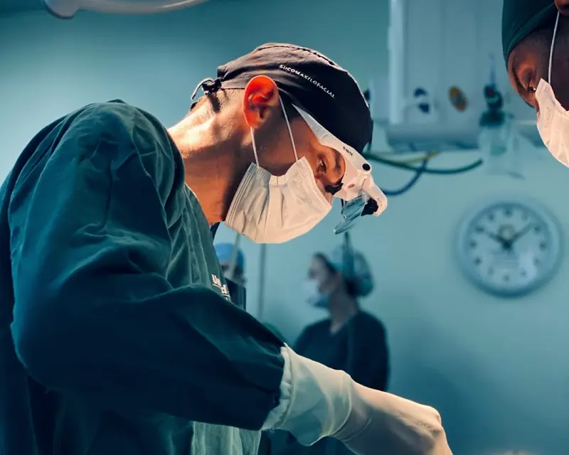 Dr. Samir Veran operando, com lupa cirúrgica e gorro de buco-maxilo-facial